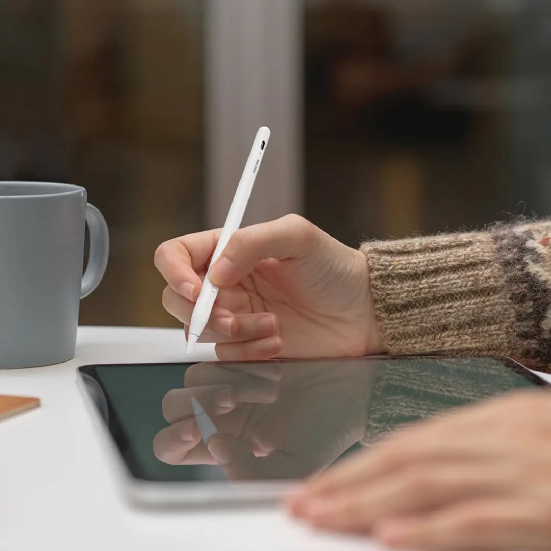 قلم لمسی سفید Pencil X در دست یک نفر، در حال نوشتن روی صفحه یک تبلت روی میز؛ کف دست دیگر روی صفحه قرار گرفته است.
