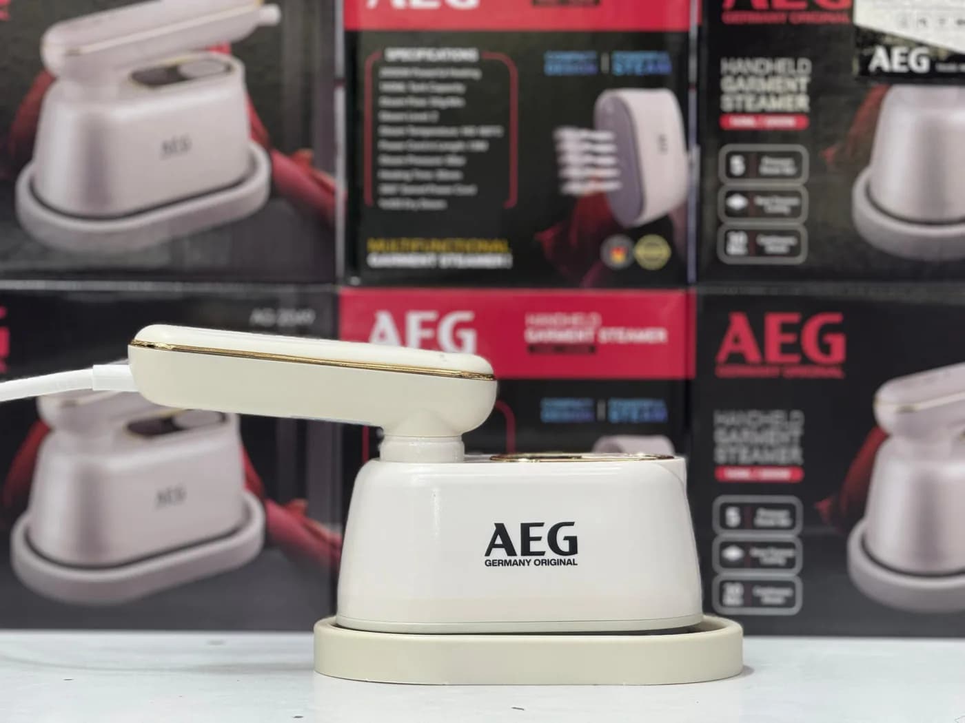 عکس واقعی بخارگر دستی AEG AG-2049 روی پایه در قفسه فروشگاه کنار جعبه‌های محصول