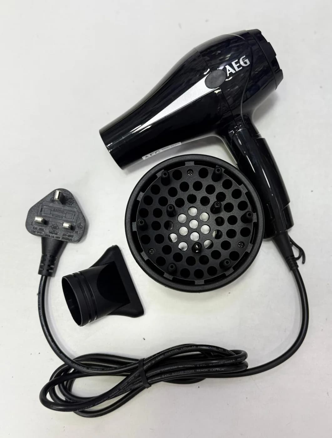 سشوار مسافرتی AEG AG-2038 به همراه دیفیوزر، نازل متمرکزکننده و کابل برق جمع‌شده با دوشاخه سه‌پین انگلیسی، روی سطح روشن